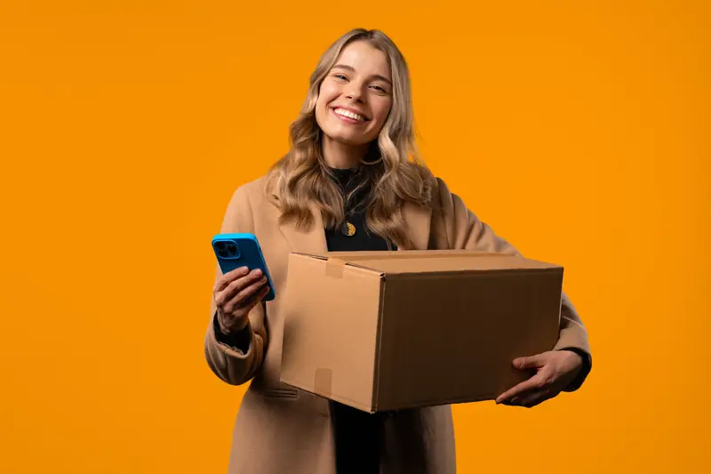 Lächelnde Frau hält ein Paket im Arm und ihr Smartphone in der Hand vor orangefarbenem Hintergrund