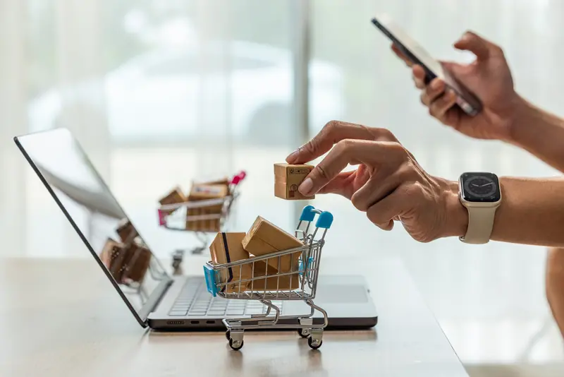 Hand legt ein kleines Paket in einen Mini-Einkaufswagen vor dem Laptop, in der anderen Hand ein Smartphone